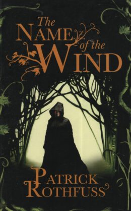 The Name of the Wind (Patrick Rothfuss po angielsku) Postać w pelerynie stoi w lesie we mgle; tytuł ‘The Name of the Wind’ widoczny u góry w ozdobnej czcionce.